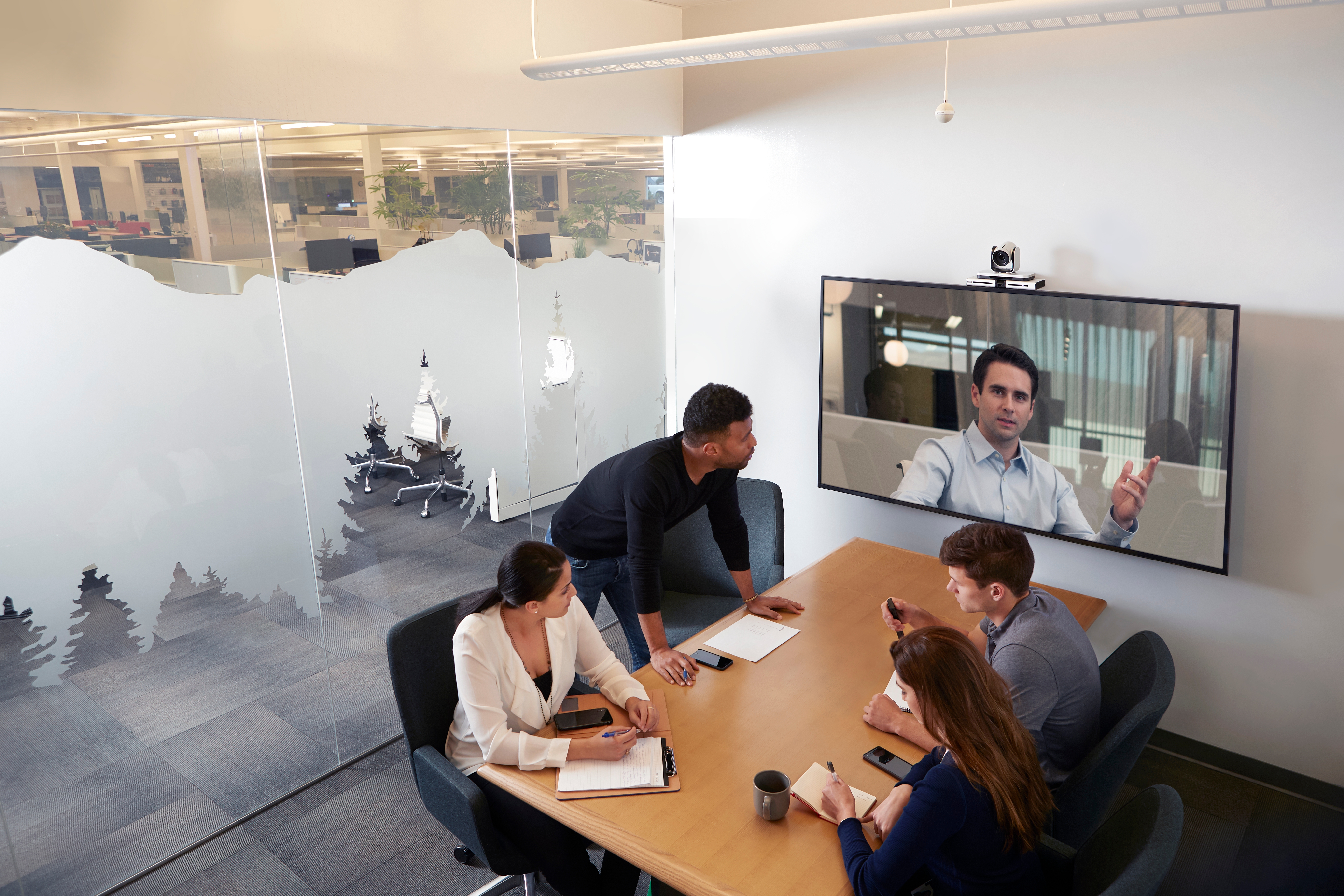 A group of four employees conducting a meeting in a conference room with a fifth employee using a video conferencing tool to join the meeting and display his camera on the TV.